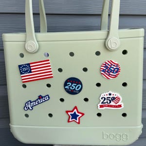 Puede incluir: Un bolso tote de color verde claro adornado con varios alfileres decorativos de temática patriótica. Los alfileres incluyen una bandera estadounidense, una estrella roja y azul y alfileres redondos con el número 250 y la palabra "America".