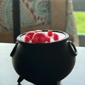May include: A black cauldron with three legs, filled with a red, bubbling liquid and several red, spherical shapes. The cauldron has two handles on the sides. The image is taken in natural light.