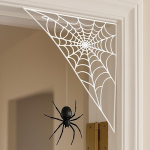 May include: White, triangular spider web decoration with a black spider hanging below. The web is mounted on a door frame, creating a spooky Halloween display. The spider has eight legs and a round body.