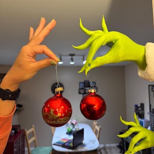 Könnte beinhalten: Zwei rote Weihnachtskugeln werden von einer menschlichen Hand und einer grünen, grinch-ähnlichen Hand gehalten. Die Kugeln haben ein glänzendes Finish und goldene Akzente. Eine Kugel hat ein goldenes Silhouettendesign. Der Hintergrund ist ein verschwommenes Interieur.