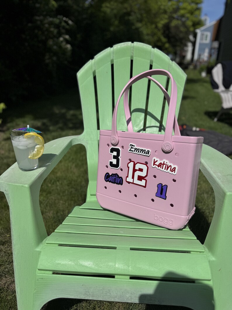 May include: A light pink tote bag with a handle sits on a green Adirondack chair. The bag has several stickers with names and numbers. A glass of lemonade with a lemon slice and a small umbrella is on the chair's arm.