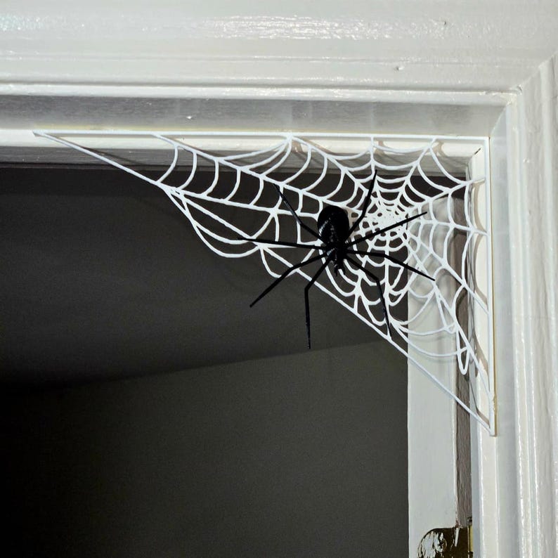 K&ouml;nnte beinhalten: Eckmontierte Spinnennetzdekoration in Wei&szlig; mit einer gro&szlig;en schwarzen Spinne. Das Netz besteht aus einem d&uuml;nnen, ausgeschnittenen Material und befindet sich an einem wei&szlig;en T&uuml;rrahmen, wodurch ein Halloween- oder gruseliges Thema entsteht.