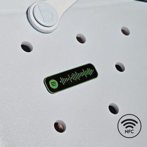 May include: Light blue shoe with a Spotify code sticker and NFC symbol. The sticker is black and green, with a barcode-like design. The shoe has several round holes and a white strap.