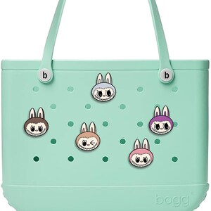Könnte beinhalten: Eine hellgrüne Tragetasche mit weißen Griffen und Aufklebern mit Cartoon-Hasengesichtern. Auf der Tasche ist der Markenname "bogg" auf der Unterseite aufgedruckt. Die Hasengesichter haben unterschiedlich farbige Ohren und Ausdrücke.