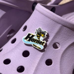 Puede incluir: Primer plano de un zueco lila con un charm decorativo. El charm presenta un avispón amarillo y negro con la palabra "Hornet" en letras negras y doradas, y "Swim Club" en letras blancas.