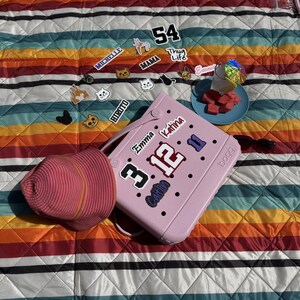 May include: A pink tote bag with various decorative stickers, including the numbers "3" and "12", and the names "Emma" and "Katina". A pink sun hat rests beside the bag on a striped, colorful blanket. A small plate of watermelon and a drink are also on the blanket.