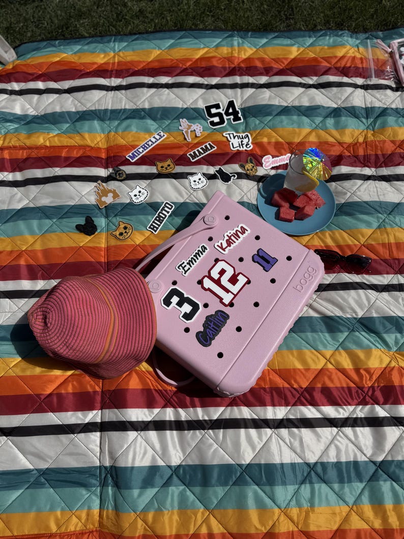 May include: A pink tote bag with various decorative stickers, including the numbers "3" and "12", and the names "Emma" and "Katina". A pink sun hat rests beside the bag on a striped, colorful blanket. A small plate of watermelon and a drink are also on the blanket.
