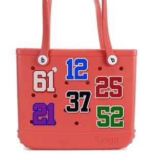May include: A coral-colored tote bag with two handles and the word "bogg" printed on the bottom. The bag features various numbers in different colors: 61, 12, 25, 37, 52, and 21. The numbers are outlined in white.