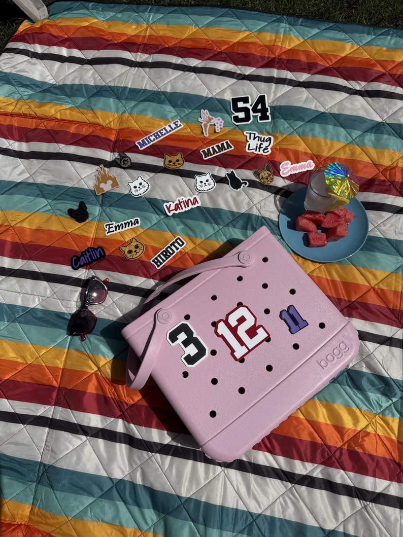 May include: A pink tote bag with the numbers "3.12" and "11" on it, surrounded by black dots. The bag is on a colorful striped blanket with various stickers and a plate of watermelon. Sunglasses are also present.
