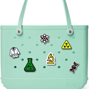 Puede incluir: Un bolso tote verde claro con asas blancas y abalorios con temática científica. Los abalorios incluyen una bata de laboratorio, un átomo, un matraz, un microscopio, una cadena de ADN y un símbolo de radiación. La bolsa tiene la palabra "bogg".