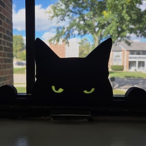 May include: A black cat silhouette with glowing green eyes is positioned in a window. The cat's head and paws are visible, creating a playful, decorative element. The background shows a blue sky, trees, and a building.