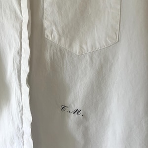 May include: A close-up of a white shirt with a pocket on the chest. The shirt has a classic collar and a button-up front. The initials "C.M." are embroidered in black on the lower right side of the shirt.