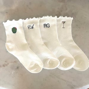 Puede incluir: Cuatro pares de calcetines blancos acanalados con volantes. Cada calcetín presenta un diseño bordado diferente: una aceituna, las letras "CA", las letras "RG" y una copa de martini. Los calcetines están colocados sobre una superficie clara.