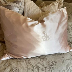 Puede incluir: Una almohada de satén rosa pálido con un monograma bordado. La almohada tiene un brillo sutil y una forma rectangular. El monograma está en un estilo ornamentado, agregando un toque de elegancia a la ropa de cama.