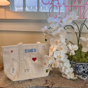 May include: A cream-colored canvas tote bag featuring embroidered icons including a red cherry, a white swan, a blue seahorse, two martini glasses, a red heart with NYC text, a blue bow, and a slice of pizza. The word CHES is embroidered in blue.