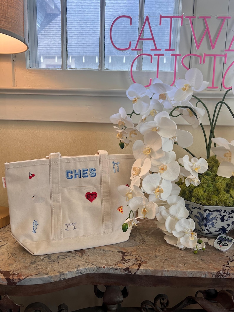 May include: A cream-colored canvas tote bag featuring embroidered icons including a red cherry, a white swan, a blue seahorse, two martini glasses, a red heart with NYC text, a blue bow, and a slice of pizza. The word CHES is embroidered in blue.