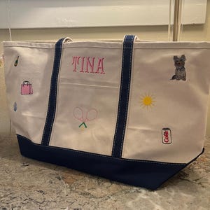 May include: A cream-colored canvas tote bag with navy blue trim and handles. The bag features embroidered icons including a champagne bottle, pink handbag, tennis rackets, a yellow sun, a dog, and a soda can with the text Coke. The name TINA is embroidered in pink.