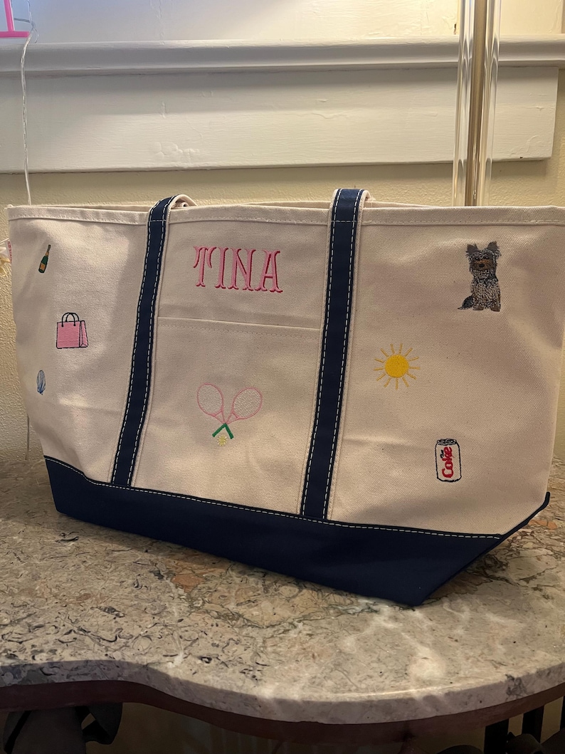 May include: A cream-colored canvas tote bag with navy blue trim and handles. The bag features embroidered icons including a champagne bottle, pink handbag, tennis rackets, a yellow sun, a dog, and a soda can with the text Coke. The name TINA is embroidered in pink.
