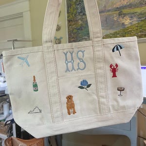 May include: A cream-colored canvas tote bag featuring embroidered designs including the letters HS, a light blue airplane, a champagne bottle, a map outline, a golden dog, a blue hydrangea, a black umbrella, a red lobster, and a cocktail glass.