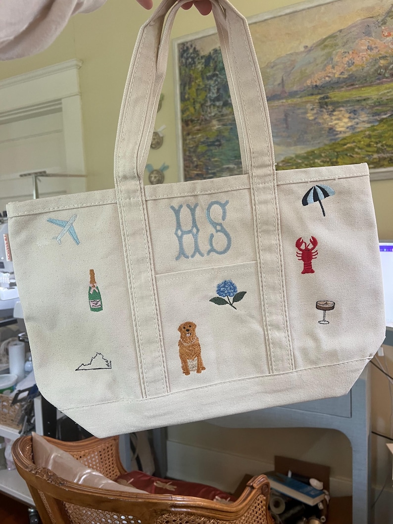 May include: A cream-colored canvas tote bag featuring embroidered designs including the letters HS, a light blue airplane, a champagne bottle, a map outline, a golden dog, a blue hydrangea, a black umbrella, a red lobster, and a cocktail glass.