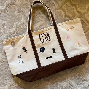 May include: A cream-colored canvas tote bag with brown trim and handles. The bag features embroidered icons including a dog, olives, a baseball cap, a lighter, sunglasses, a bit, a spool of thread, and lipstick. The monogram CM is centered.
