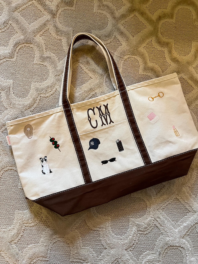 May include: A cream-colored canvas tote bag with brown trim and handles. The bag features embroidered icons including a dog, olives, a baseball cap, a lighter, sunglasses, a bit, a spool of thread, and lipstick. The monogram CM is centered.