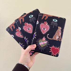 May include: Four black covers for an e-reader with a repeating pattern of hearts, eyes, and leopards. The hearts are red and pink with an eye in the center. The eyes are green. The leopards are orange and black.