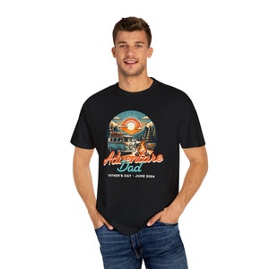 Puede incluir: Una camiseta negra con una ilustración de estilo retro de una furgoneta de camping, montañas y una fogata. El texto "Adventure Dad" está escrito en una fuente estilizada, con las palabras "Father's Day - June 2024" debajo.