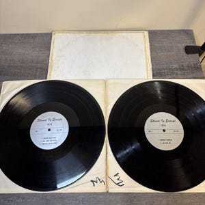 Puede incluir: Dos discos de vinilo negros del álbum "Stoned In Europe 1970" se muestran en sus fundas de papel. La portada del álbum es visible en el fondo. Las etiquetas de los discos son blancas con texto negro.