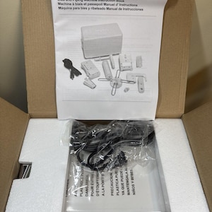 May include: A Simplicity Bias and Piping Machine instruction book with a diagram of parts, including a white machine, power cord, and various attachments. The manual is placed inside a cardboard box with white foam packaging and a plastic-wrapped item.