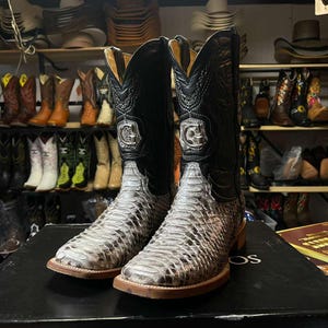 Könnte beinhalten: Ein Paar Cowboy-Stiefel mit Schlangenmuster am unteren Teil und schwarzen Lederoberteilen. Die Stiefel haben ein silbernes Emblem und eine braune Sohle. Weitere Stiefel sind auf Regalen im Hintergrund zu sehen.