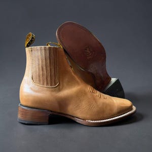 May include: A pair of tan leather ankle boots with a square toe and stacked heel. One boot is upright, showcasing the side and front details, while the other is inverted, revealing the sole. The boots have a classic Western design.