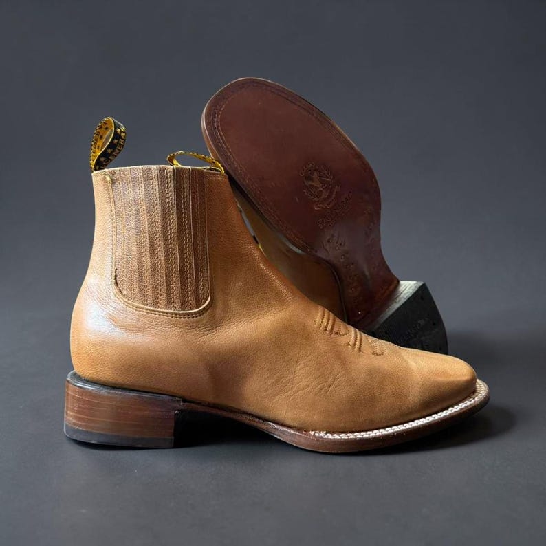 May include: A pair of tan leather ankle boots with a square toe and stacked heel. One boot is upright, showcasing the side and front details, while the other is inverted, revealing the sole. The boots have a classic Western design.