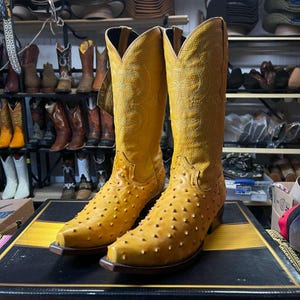 May include: A pair of yellow cowboy boots with a textured, studded design on the foot and lower leg. The boots have a pointed toe and a decorative pattern on the upper shaft. Other boots are visible in the background.