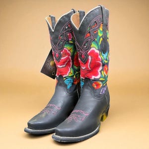 May include: A pair of black leather cowboy boots with vibrant floral designs in red, yellow, blue, and green. The boots have pink stitching and a yellow heel. A tag hangs from one boot.