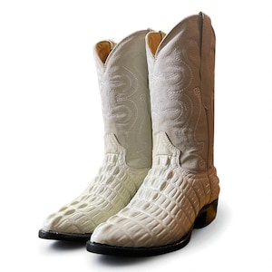 Könnte beinhalten: Ein Paar cremefarbene Cowboy-Stiefel. Die Stiefel haben ein strukturiertes, krokodilartiges Muster auf dem Fuß und ein gesticktes Design auf dem Schaft. Die Sohlen sind schwarz und die Absätze sind dunkelbraun.