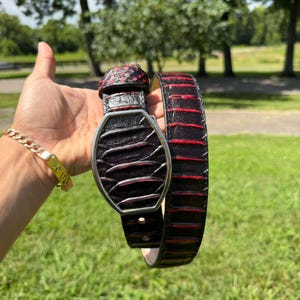May include: A black leather belt featuring a textured, scale-like pattern with deep red accents between the ridges. The belt includes a distinctive, elongated hexagonal silver-toned metal buckle that secures the strap in a looped configuration.