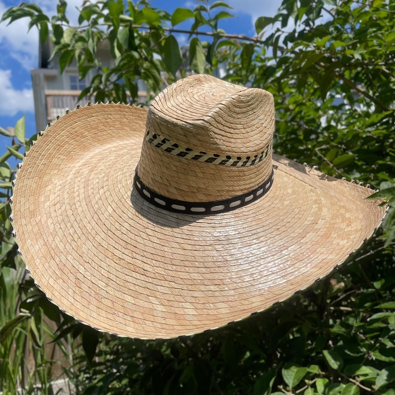 May include: A wide-brimmed straw hat with a natural beige color. The hat features a black and white band around the crown and a decorative trim around the brim. The hat is set against a backdrop of green foliage and a blue sky.