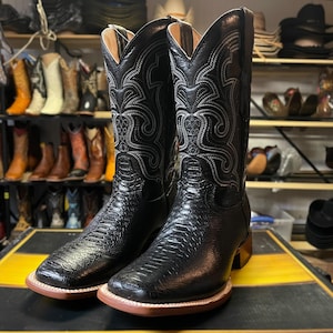 May include: A pair of black cowboy boots with a textured pattern and decorative stitching. The boots have a square toe and a brown sole. The background shows a collection of other boots and hats.