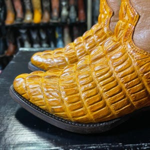 May include: Close-up of a pair of cowboy boots with a textured, yellow-orange alligator skin pattern. The boots have a brown leather upper and a dark brown sole. The boots are displayed on a black surface.