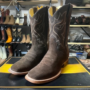 May include: A pair of brown cowboy boots with a square toe and decorative stitching. The boots have a suede-like texture on the foot and a smooth leather upper. The soles are light brown, and the interior is yellow.