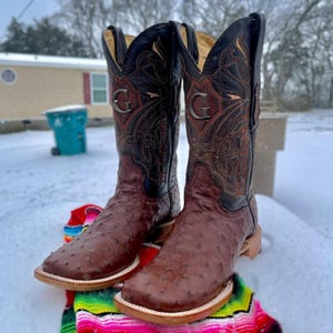 May include: A pair of brown cowboy boots with a square toe and decorative stitching. The boots have a black upper with a silver "G" emblem. The boots are set against a snowy backdrop.