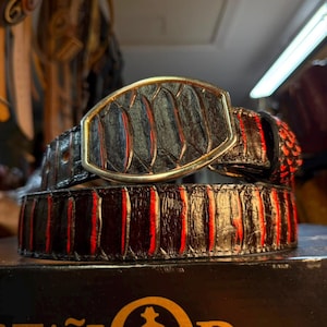 May include: A black belt featuring a textured, scale-like pattern with vibrant red accents between the raised ridges. The belt includes a large, oval-shaped buckle with a metallic gold-toned frame that matches the belt's textured material.