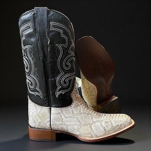May include: Cowboy boots with a black leather upper and a white snakeskin-patterned foot. The black upper has detailed white stitching. The boots have a brown sole and heel, set against a dark background. The boots are a classic Western style.