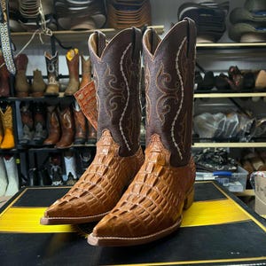 May include: A pair of brown cowboy boots with a textured, crocodile-like pattern on the lower section and a decorative stitched design on the upper shaft. The boots are displayed on a black and yellow surface, with other boots visible in the background.