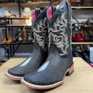 May include: A pair of black cowboy boots with gold decorative stitching. The boots have a textured finish, a light-coloured toe, and a pink interior lining. The boots are displayed on a cardboard surface.