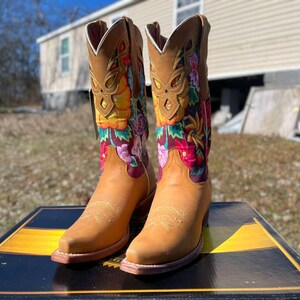 May include: Tan leather cowboy boots with vibrant floral embroidery. The boots feature a pointed toe, a stacked heel, and a tall shaft. The floral design incorporates red, yellow, and pink flowers. The boots are displayed on a black and gold surface.
