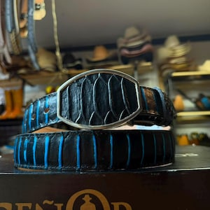 May include: A black leather belt featuring a textured, scale-like pattern with vibrant blue accents between the ridges. The belt includes a prominent, oval-shaped silver-toned metal buckle that frames the textured leather material at the center.