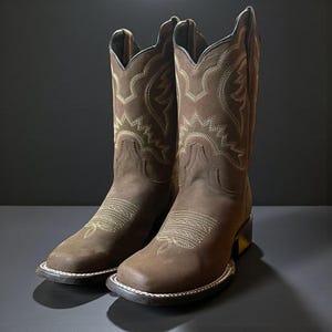 May include: A pair of brown cowboy boots with cream-coloured decorative stitching. The boots have a square toe and a stacked heel. The leather has a matte finish. The boots are displayed against a dark background.