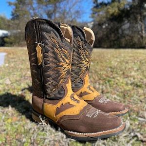 May include: A pair of brown leather cowboy boots with detailed stitching and a square toe. The boots feature dark brown, tan, and black leather, with gold and white decorative stitching. The boots are designed in a Western style.